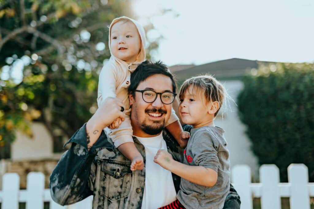 A man holding his two children in front of a white picket.