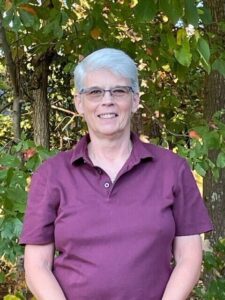 A woman in a maroon shirt standing in front of trees.