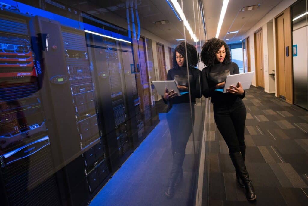 A woman is standing in a server room looking at a laptop.
