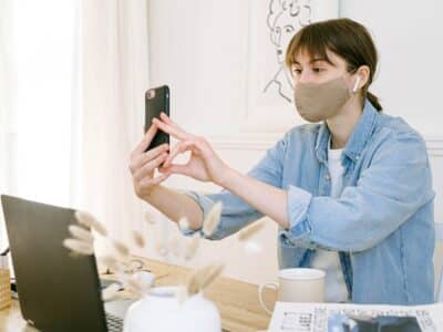 A woman wearing a face mask is taking a picture of her laptop.