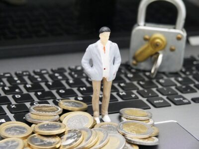 A toy man standing on a pile of coins next to a laptop.