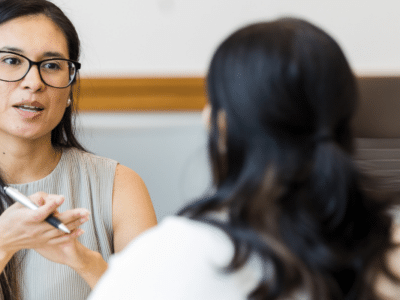 Two women in glasses talking to each other in a conference room.