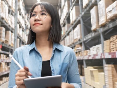 A woman is standing in a warehouse holding a clipboard.