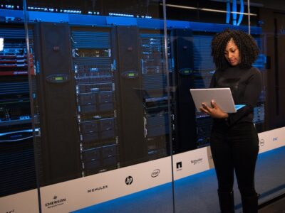 A woman standing in front of a server room with a laptop.