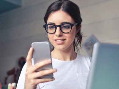 A woman in glasses is looking at her phone while sitting at a desk.