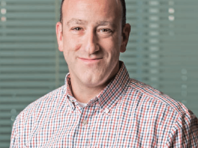 A man in a checkered shirt smiling in front of a window.