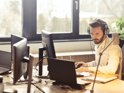 A man sitting at a desk with two computers and a headset.