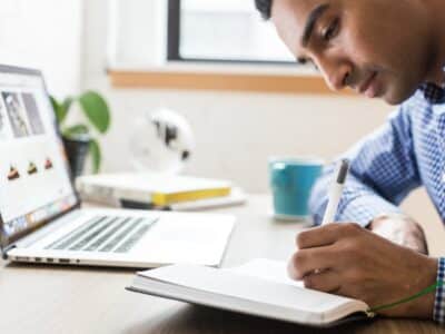 A man writing in a notebook while sitting at a desk with a laptop.