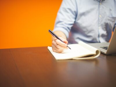 A man writing on a notebook with a pen in front of an orange background.