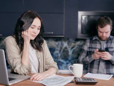 A man and woman are sitting at a kitchen table while looking at their cell phones.