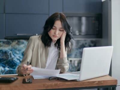 A woman sitting at a kitchen table with a laptop and papers.