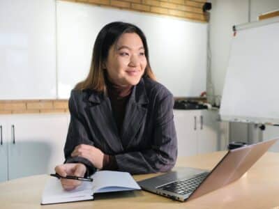 A young asian woman sitting at a desk with a notebook and laptop.