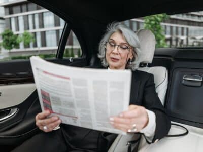 A woman is sitting in the back seat of a car reading a newspaper.