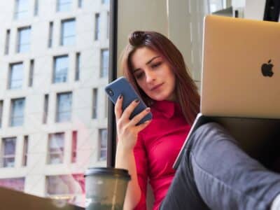 A woman sitting in front of a window with a laptop and cell phone.