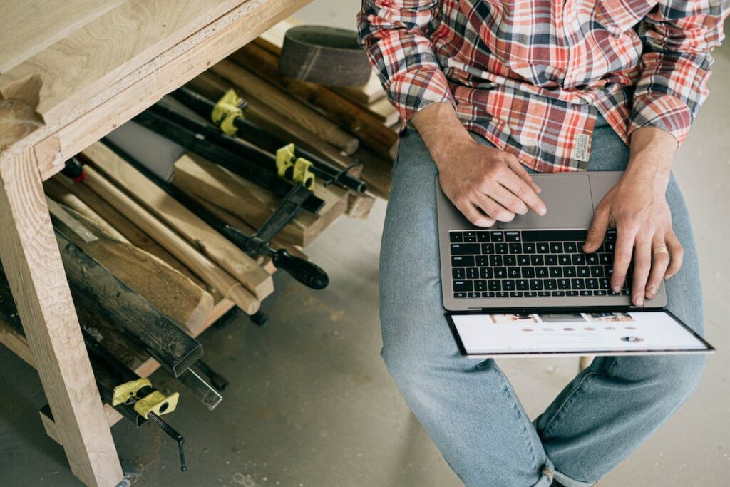 A man at a construction site using accounting software on a laptop and wearing a plaid shirt with blue jeans.