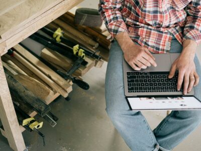A man at a construction site using accounting software on a laptop and wearing a plaid shirt with blue jeans.