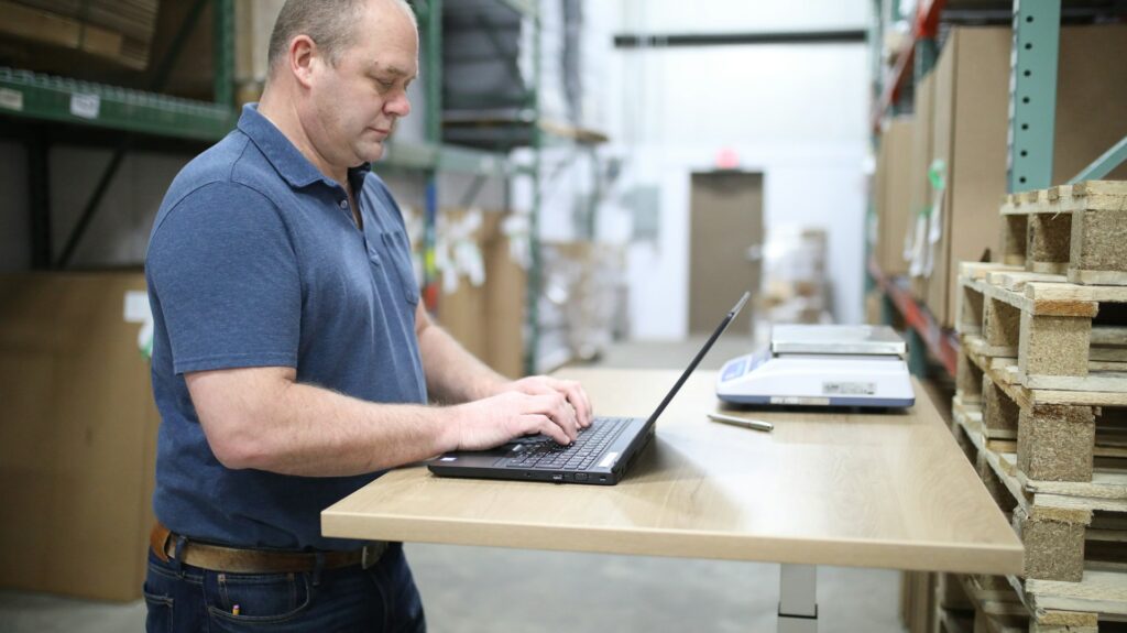 White male standing at a desk using accounting software on his laptop in a manufacturing facility.