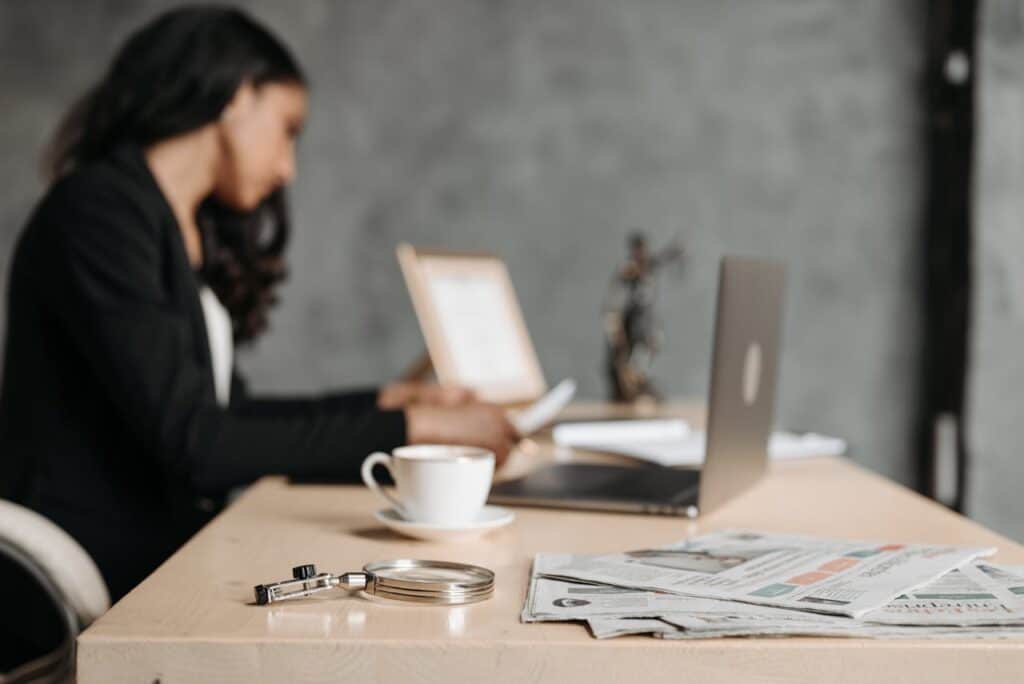 An out of focus shot of a woman in a black suit sitting at a desk looking reports while additional reports and her laptop are in the background, as she learns to drill down in Generic Inquiries in Acumatica