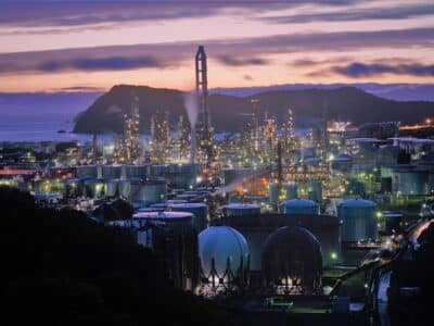 A smart factory complex with over a dozen silos and a large tower in the middle at night next to a bay and several hills overlooking the facility, with bright lights all over the structures.