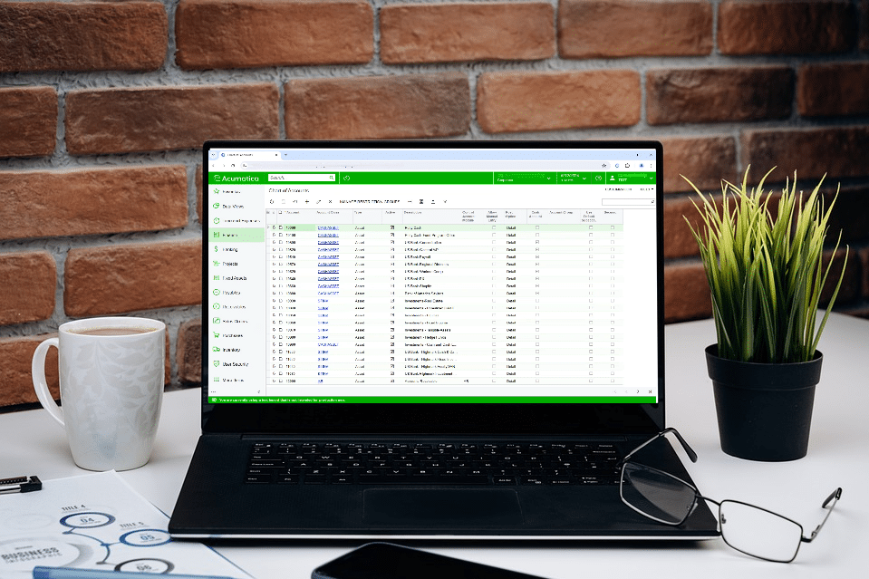 Black laptop displaying Acumatica General Ledger journal transactions screen on a desk with coffee mug and notepad, showcasing the financial management interface on a computer in an office setting.