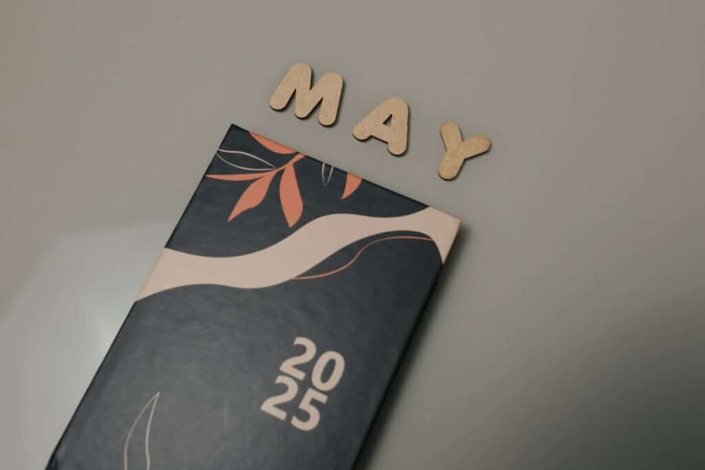 Wooden letters spelling "MAY" above a black 2025 calendar page with decorative coral floral design on gray background