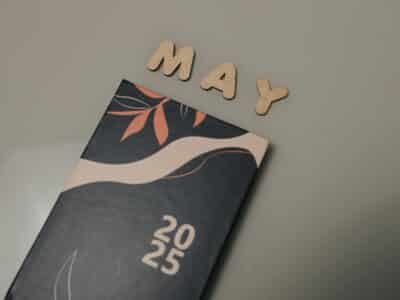 Wooden letters spelling "MAY" above a black 2025 calendar page with decorative coral floral design on gray background