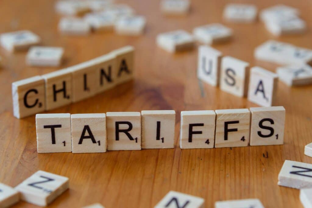 Wooden letter tiles spelling "TARIFFS" in the foreground with "CHINA" and "USA" visible in the background, illustrating trade tensions affecting technology hardware costs.