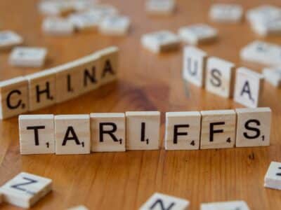 Wooden letter tiles spelling "TARIFFS" in the foreground with "CHINA" and "USA" visible in the background, illustrating trade tensions affecting technology hardware costs.