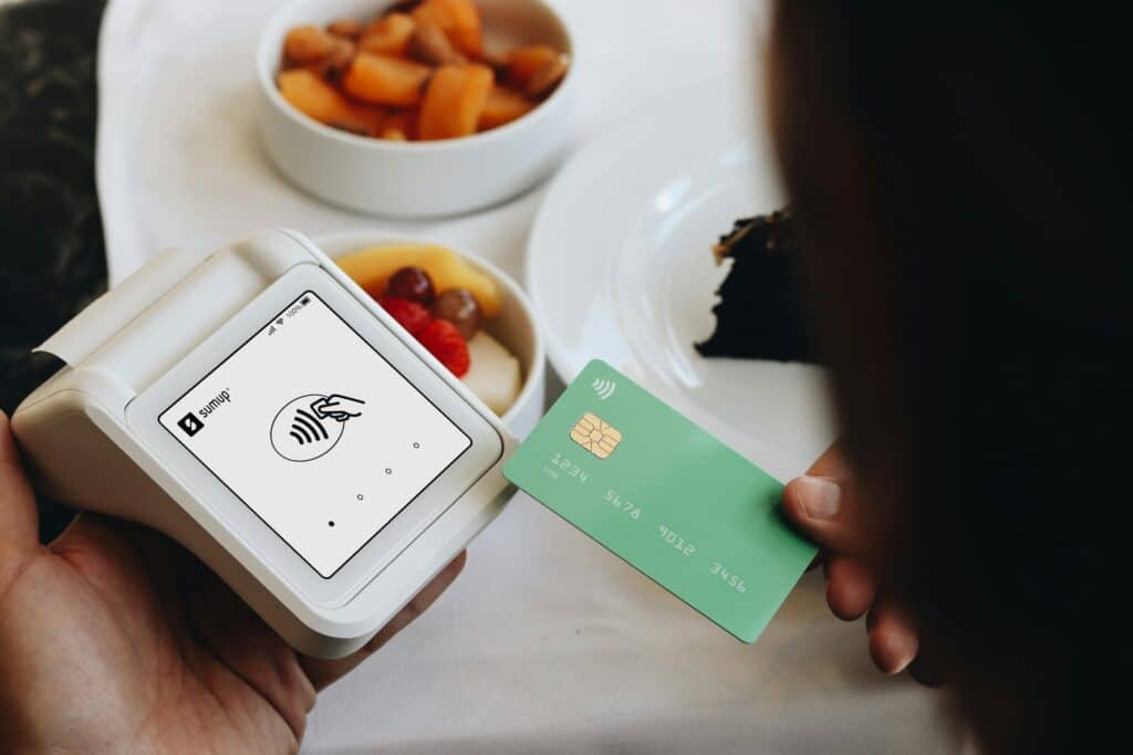 A person holding a green credit card near a white contactless payment terminal displaying wireless payment symbols, with bowls of food and a mug visible on a table in the background.