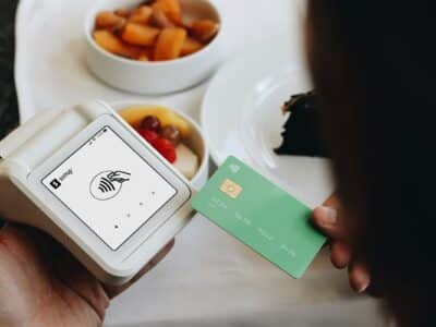 A person holding a green credit card near a white contactless payment terminal displaying wireless payment symbols, with bowls of food and a mug visible on a table in the background.
