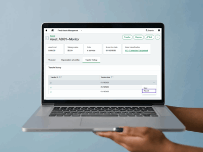 Sage Intacct Fixed Assets Management interface updated with 2025 R2 and displaying asset details including cost, depreciation schedules, and transfer history on a laptop screen being held up by a pair of hands from the right of the screen.
