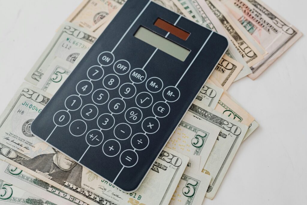 Black digital calculator with numeric keypad and function buttons positioned on top of scattered U.S. dollar bills of various denominations, representing financial calculations and accounting.