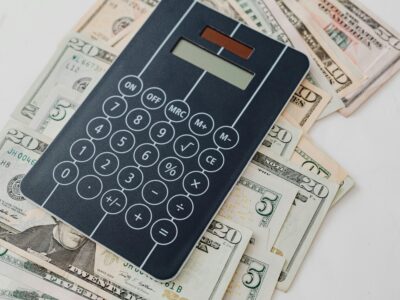 Black digital calculator with numeric keypad and function buttons positioned on top of scattered U.S. dollar bills of various denominations, representing financial calculations and accounting.
