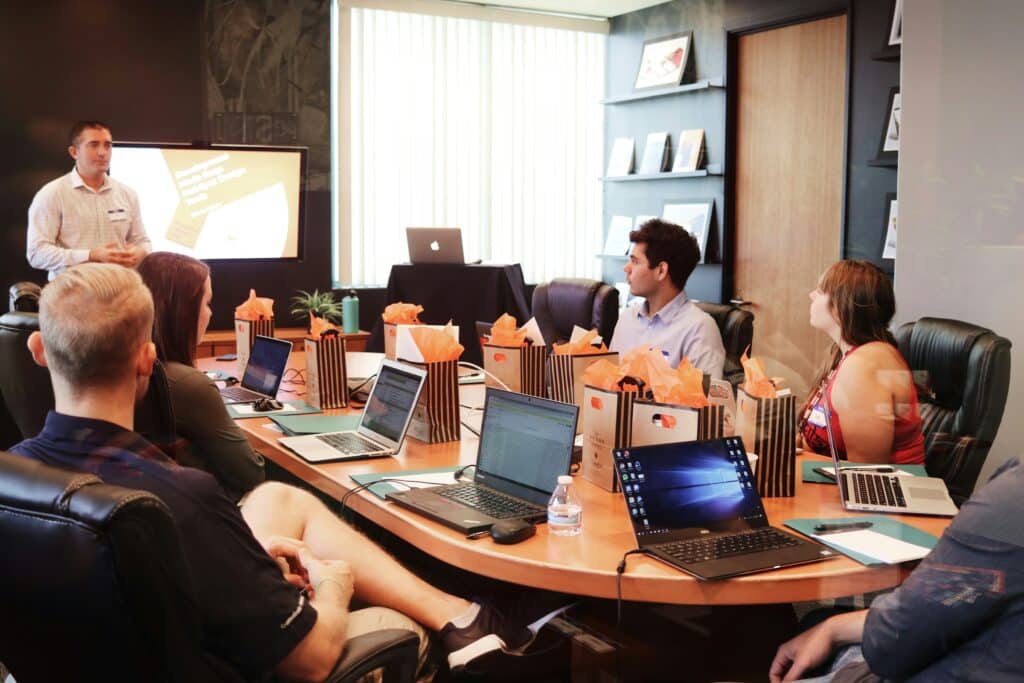 Business meeting in a modern conference room with a presenter standing near a large wall-mounted screen while several attendees sit around a wooden table with open laptops and orange gift bags.