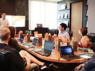 Business meeting in a modern conference room with a presenter standing near a large wall-mounted screen while several attendees sit around a wooden table with open laptops and orange gift bags.