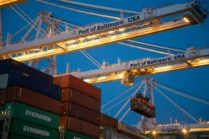 Large white container cranes at Port of Baltimore loading shipping containers including Evergreen and COSCO freight at dusk with illuminated port operations.