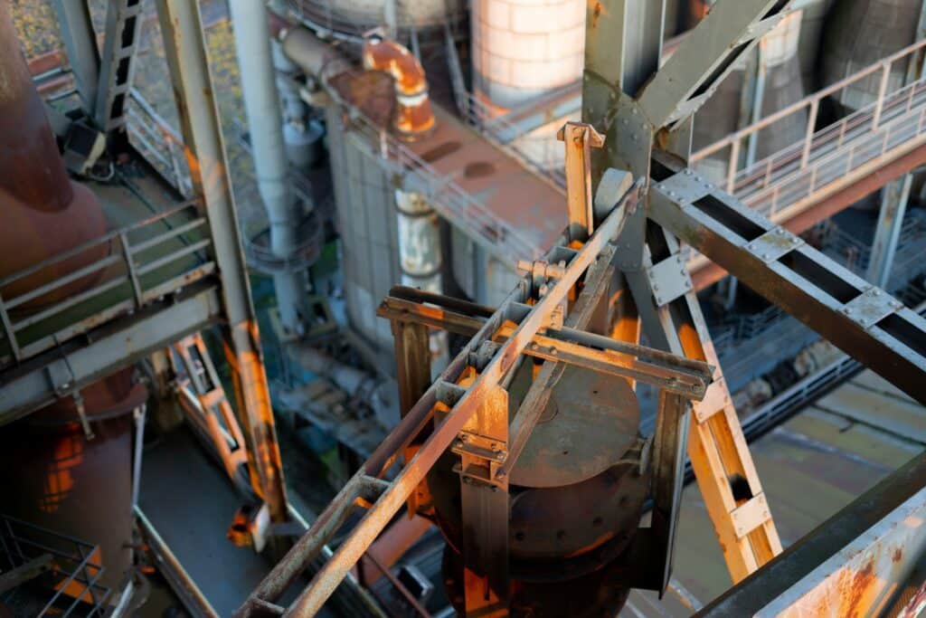 Industrial manufacturing equipment with metal and wooden components illuminated by warm lighting inside a factory facility, showing mechanical apparatus surrounded by metal railings and industrial infrastructure.