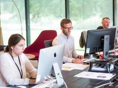 Professional services team members working on laptops and desktop computers in a modern office environment with large windows, representing collaborative work across multiple systems and platforms.