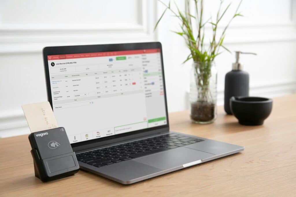 Laptop displaying business software interface on wooden desk with card payment terminal, plant in glass vase, and coffee cups in modern office setting.