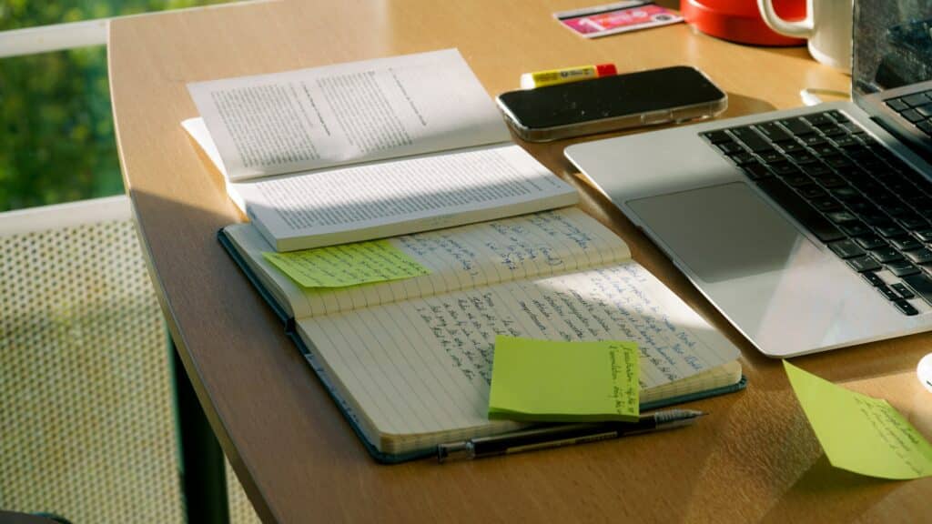 A wooden desk bathed in natural light displays an organized workspace with open notebooks containing handwritten notes, bright yellow sticky notes, printed documents, a laptop, smartphone, and red coffee mug, suggesting an active study or work session.