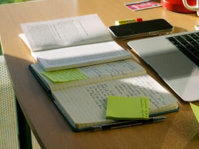A wooden desk bathed in natural light displays an organized workspace with open notebooks containing handwritten notes, bright yellow sticky notes, printed documents, a laptop, smartphone, and red coffee mug, suggesting an active study or work session.