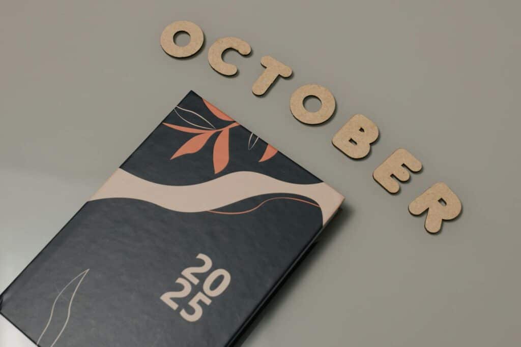 Wooden letters spelling "OCTOBER" arranged above a 2025 planner with botanical leaf design in coral and navy colors on a neutral background.