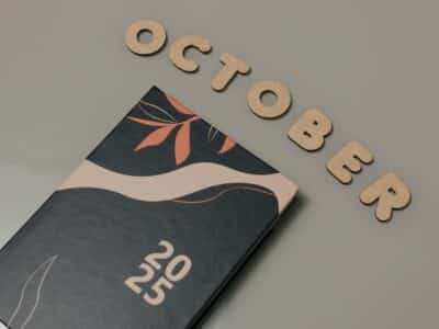 Wooden letters spelling "OCTOBER" arranged above a 2025 planner with botanical leaf design in coral and navy colors on a neutral background.