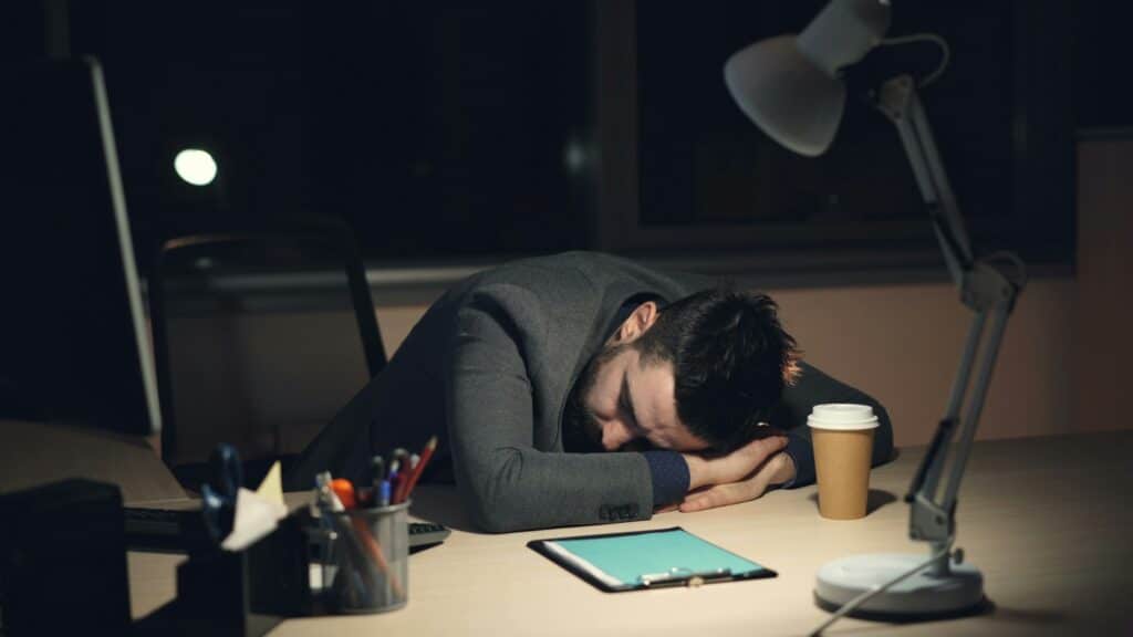 Exhausted IT professional in suit asleep at desk after hours with coffee cup, tablet showing blue screen and desk lamp, illustrating cybersecurity alert fatigue and burnout.