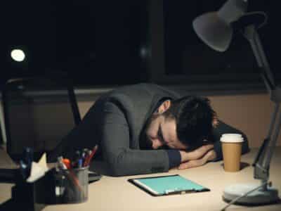 Exhausted IT professional in suit asleep at desk after hours with coffee cup, tablet showing blue screen and desk lamp, illustrating cybersecurity alert fatigue and burnout.