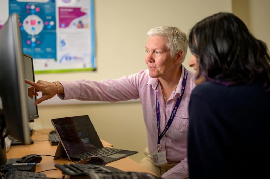 An older female IT support professional aids a female employee at a workstation, with the former on the left pointing at a desktop monitor while reviewing system configurations on a laptop.
