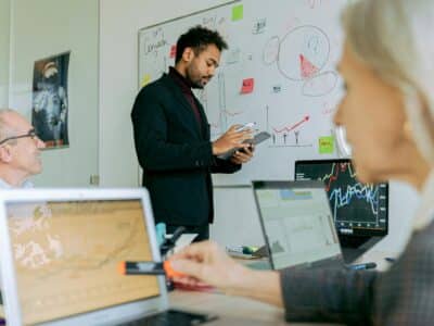 Person in a dark blazer standing at a whiteboard holding a tablet, with two people seated at a table with open laptops in the foreground appearing to take notes while the former is conducting cybersecurity training.
