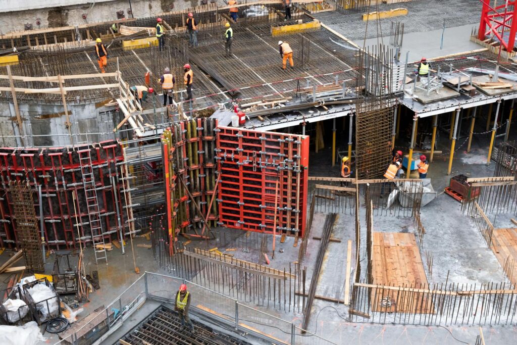 Aerial view of an active construction jobsite with multiple workers in orange safety vests and hard hats working across concrete formwork, rebar grids and red metal shoring panels during a large-scale building project.