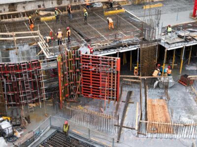 Aerial view of an active construction jobsite with multiple workers in orange safety vests and hard hats working across concrete formwork, rebar grids and red metal shoring panels during a large-scale building project.