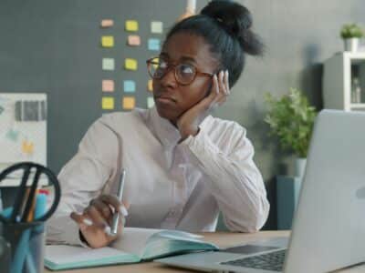 Woman wearing glasses and an off-white button-down shirt sitting at a desk with a laptop, open notebook, and pen, resting their cheek on their hand while looking away in thought, with colorful sticky notes on a gray wall in the background.